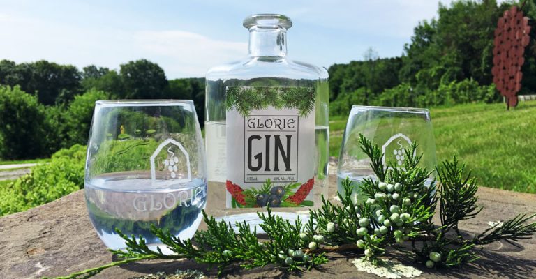 Bottle of Glorie Farm Gin with two glasses and juniper berries outdoors on a rock.