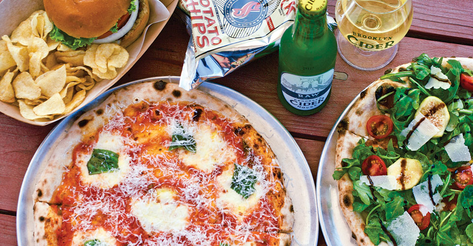 plated pizza and burgers on a picnic table with a bottle of Brooklyn Cider