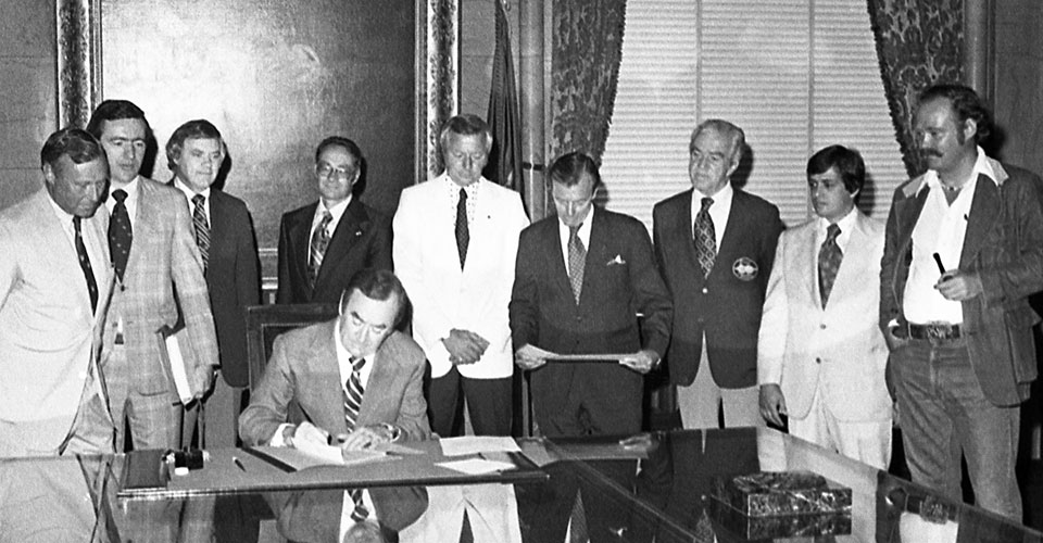 b/w photo of Gov Hugh carey signs document with group of men standing behind